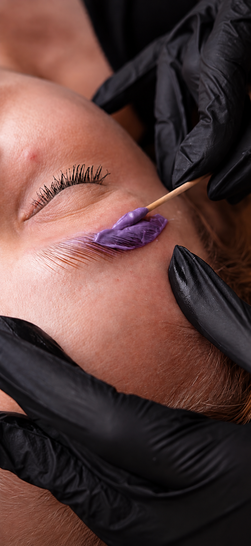 Profissional de luvas pretas aplicando o produto roxo da PRISME na sobrancelha com espátula